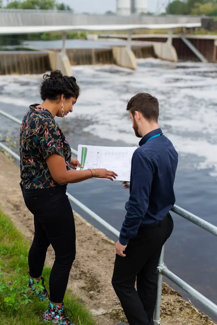 Male and female engineers reviewing plans outdoors by a river dam.